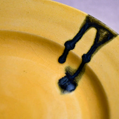Close up of a yellow ceramic dinner plate showing black drips, texture, and crazing.