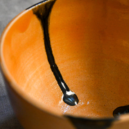 Close-up of a ceramic bowl interior with a warm orange glaze and a bold black painted accent running diagonally across the surface.