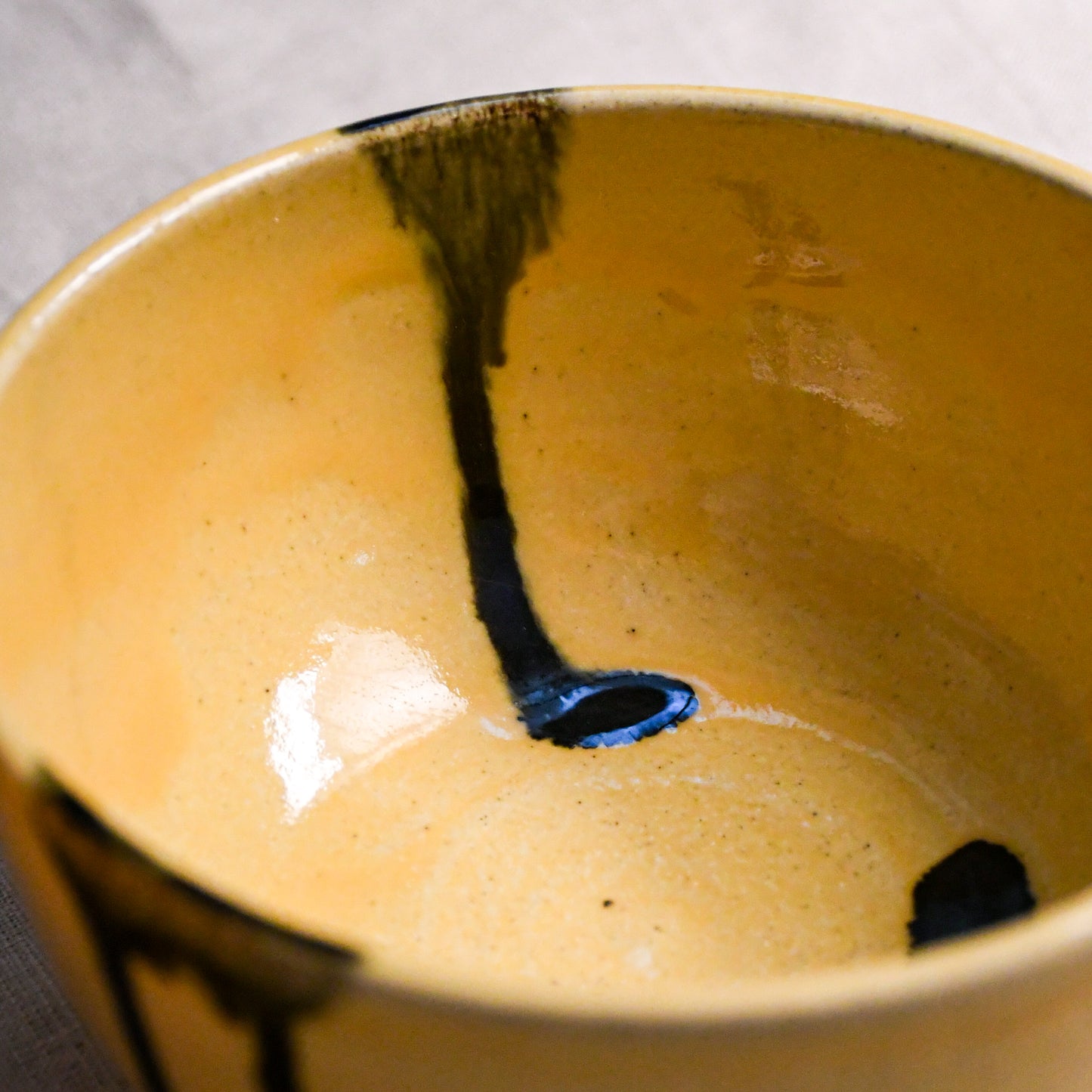 Close-up of a ceramic bowl with a glossy mustard-yellow interior and dark brown to black abstract drips near the rim and base.