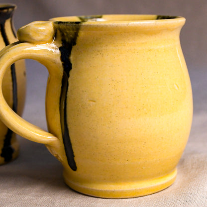 A close-up of a handmade ceramic mug with a textured yellow glaze and dark brown glaze drips, set against a neutral gray background.