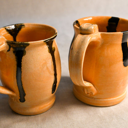 Two handmade ceramic mugs with a warm orange-peach glaze and dark brown to black dripped accents, set against a neutral beige background.