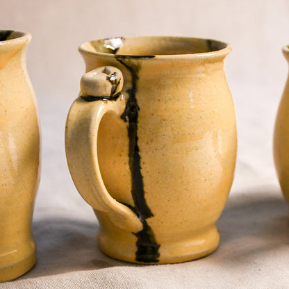 A handmade ceramic mug with a warm yellow-beige speckled glaze and a prominent dark brown vertical crack, shown in a close-up shot with other mugs partially visible on either side.