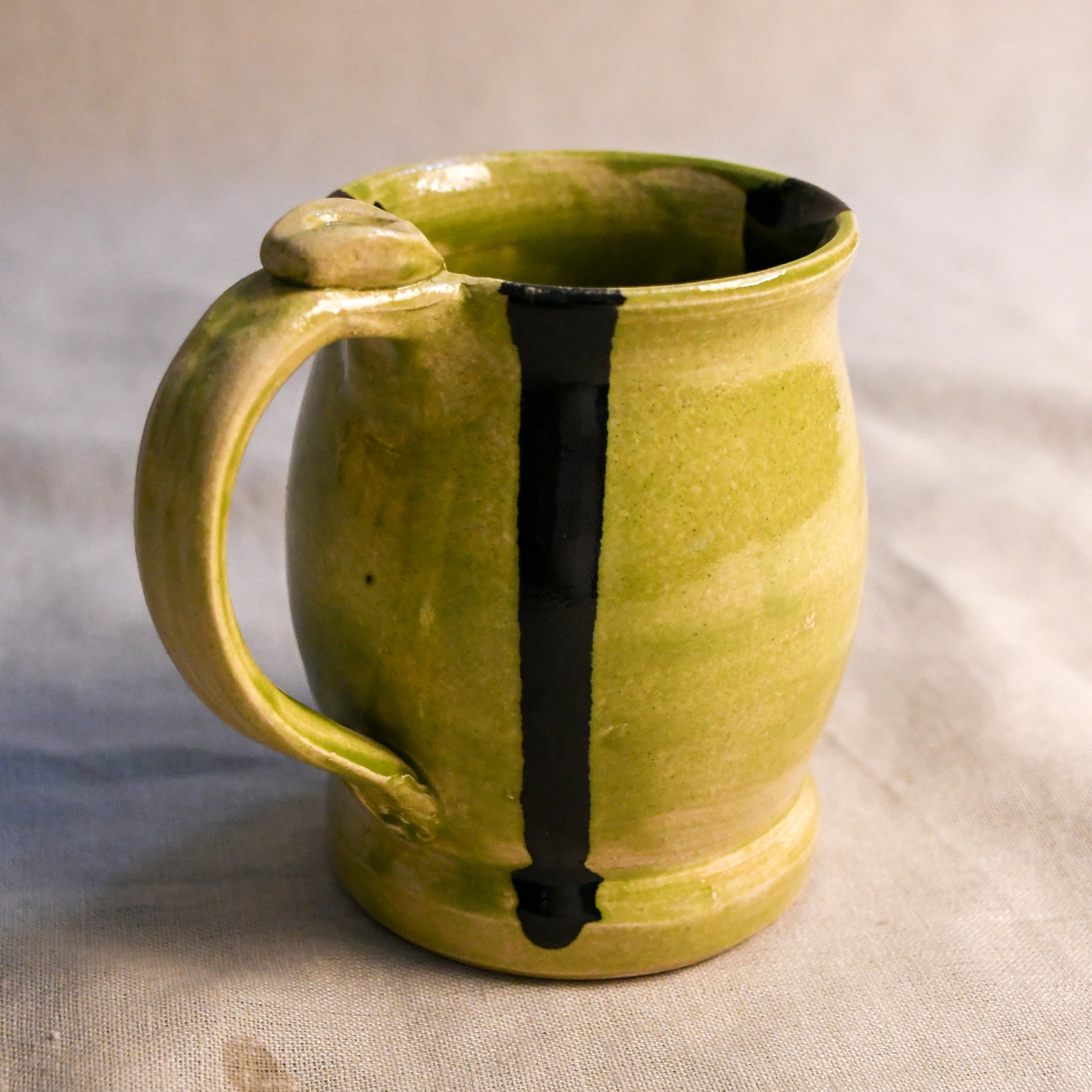 A handmade ceramic mug with a textured olive-green glaze and a vertical black stripe, set against a neutral fabric background.