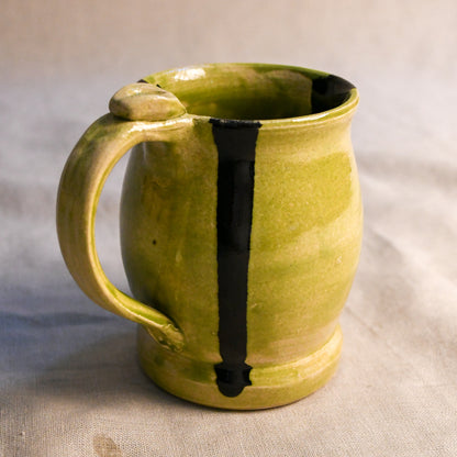 A handmade ceramic mug with a textured olive-green glaze and a vertical black stripe, set against a neutral fabric background.