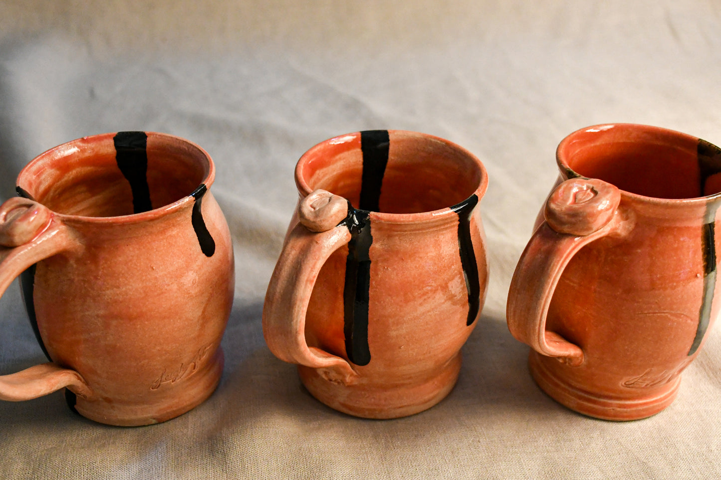 Three handmade terracotta clay mugs with black painted drips, arranged side-by-side on a neutral beige fabric background.