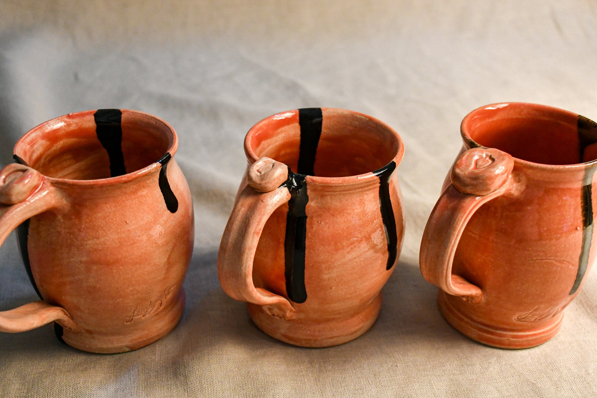 Three handmade terracotta clay mugs with black painted drips, arranged side-by-side on a neutral beige fabric background.