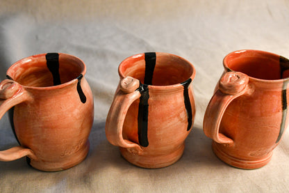 Three handmade terracotta clay mugs with black painted drips, arranged side-by-side on a neutral beige fabric background.