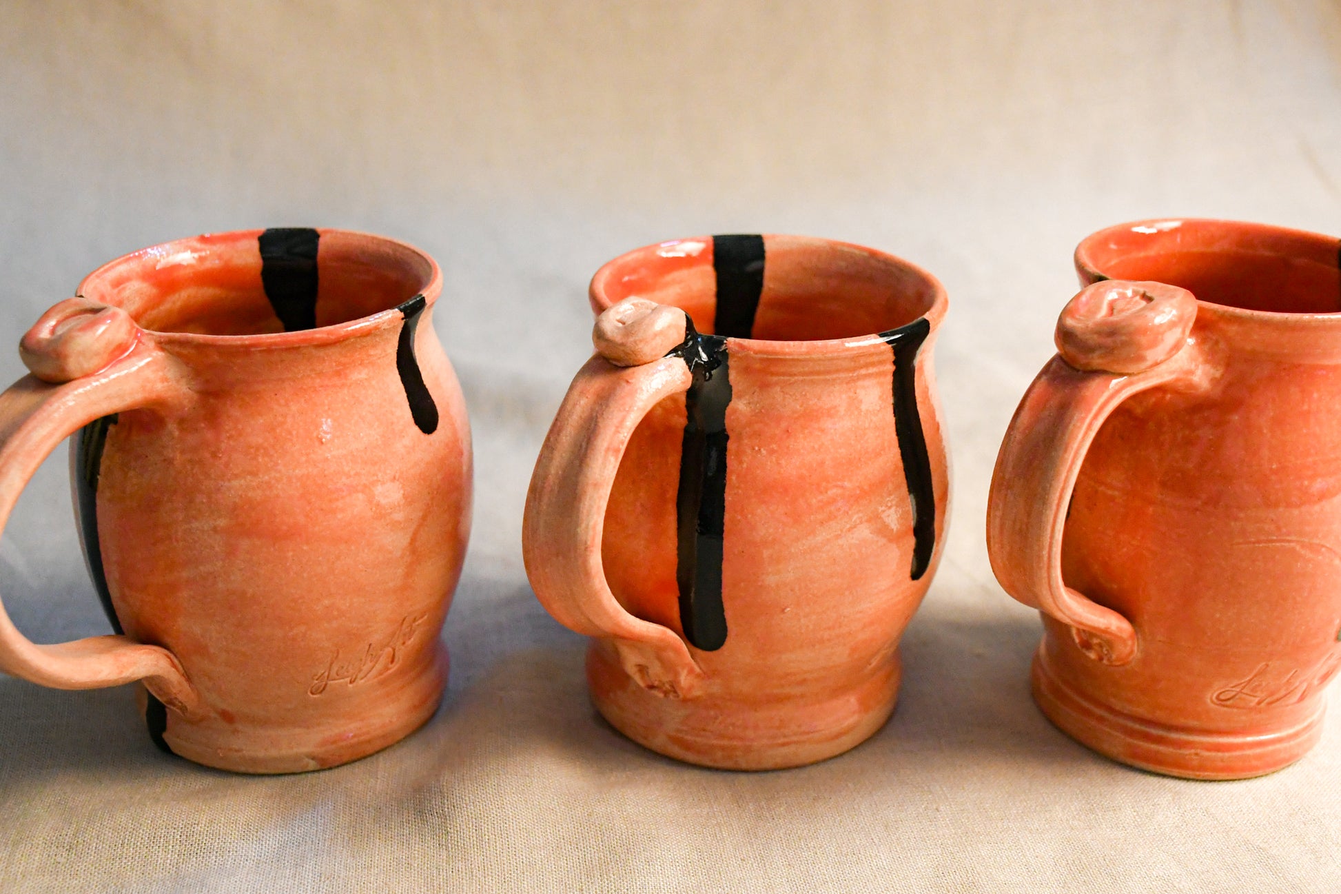 Three handmade terracotta ceramic mugs with black painted drips, arranged side-by-side on a neutral beige fabric background.