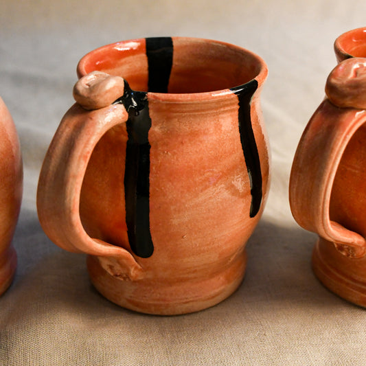 A handmade terracotta ceramic mug with a thick handle and glossy black paint drips, shown on a neutral beige fabric background.