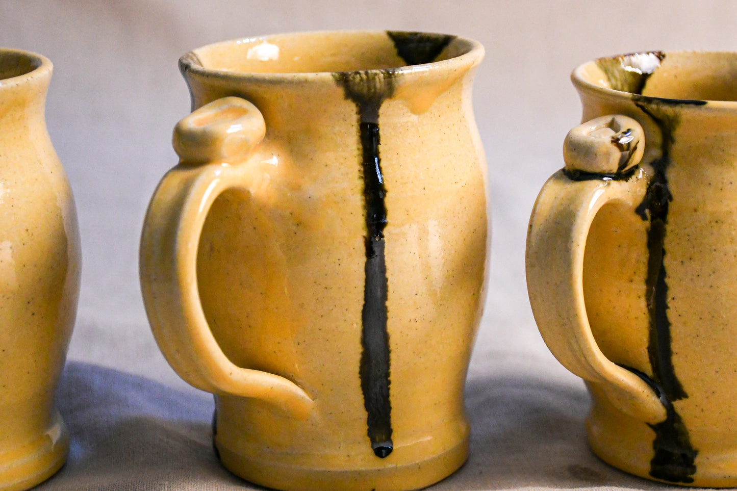 A close-up of a handmade ceramic mug with a warm yellow-beige glaze and dark brown to black vertical drips, shown alongside other partial mugs on a neutral gray background.