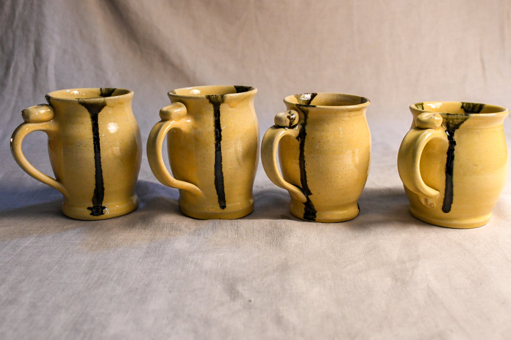 Four handmade ceramic mugs with a warm yellow glaze and dark brown vertical drips, arranged in a row against a neutral gray fabric backdrop.