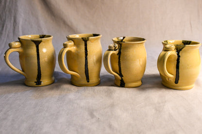 Four handmade ceramic mugs with a warm yellow glaze and dark brown vertical drips, arranged in a row against a neutral gray fabric backdrop.