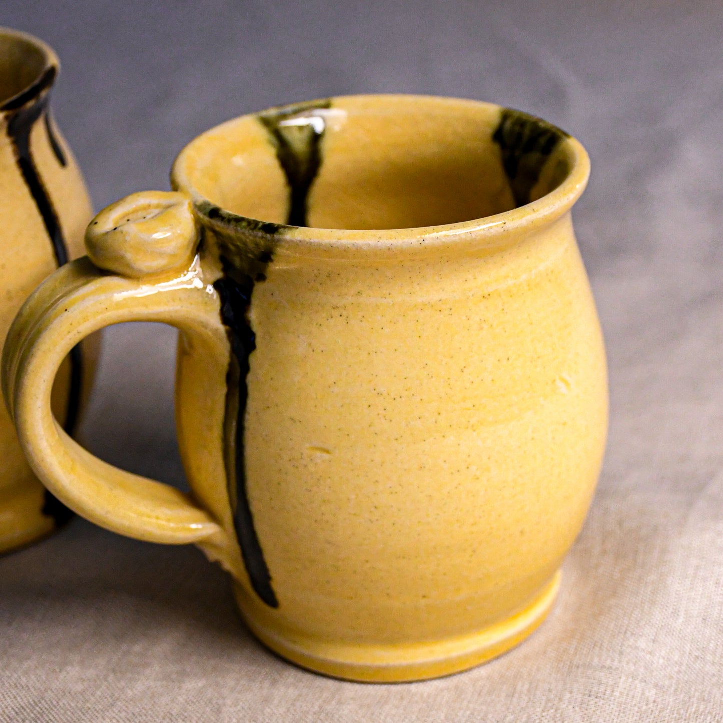 A close-up of a handmade ceramic mug with a textured yellow glaze and dark brown drips, with another mug partially visible in the background on a neutral fabric surface.