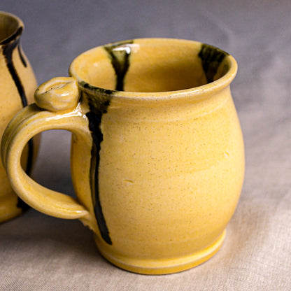 A close-up of a handmade ceramic mug with a textured yellow glaze and dark brown drips, with another mug partially visible in the background on a neutral fabric surface.