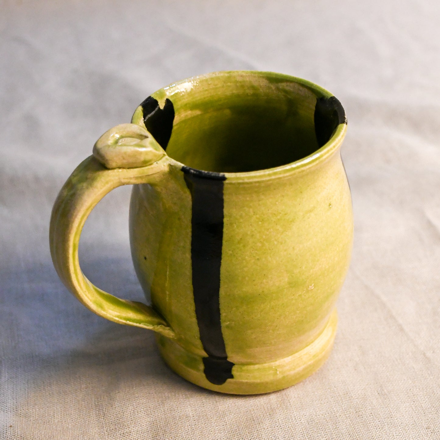 A handmade ceramic mug with a rounded body, a thick handle, and a vertical black stripe running down the center, finished in a textured olive-green glaze, set against a neutral gray fabric background.