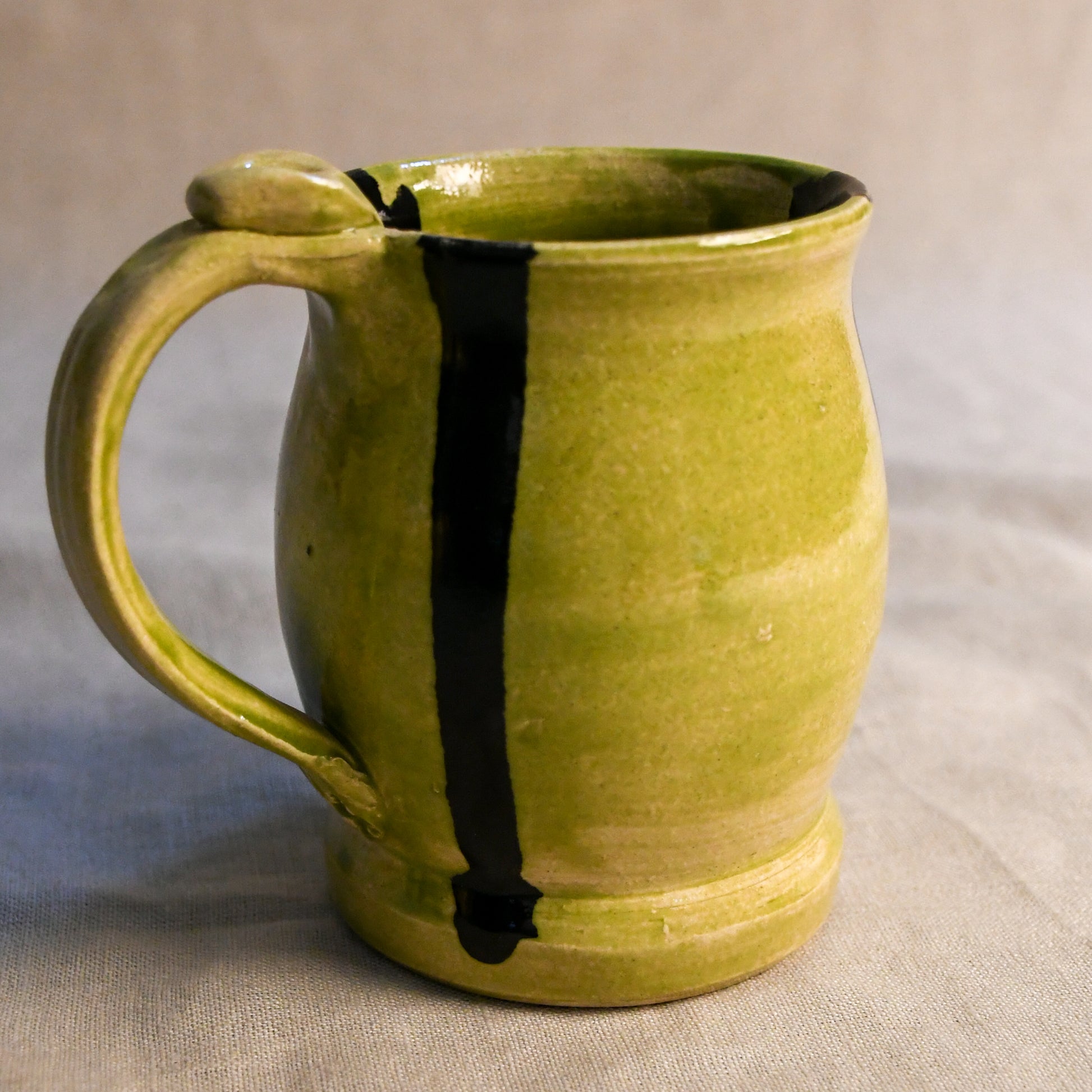A handmade ceramic mug with a textured olive-green glaze and a bold vertical black stripe, resting on a neutral gray fabric background.