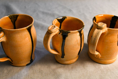 Three handmade ceramic mugs with orange-brown glaze and black dripped accents, arranged on a neutral gray fabric background.