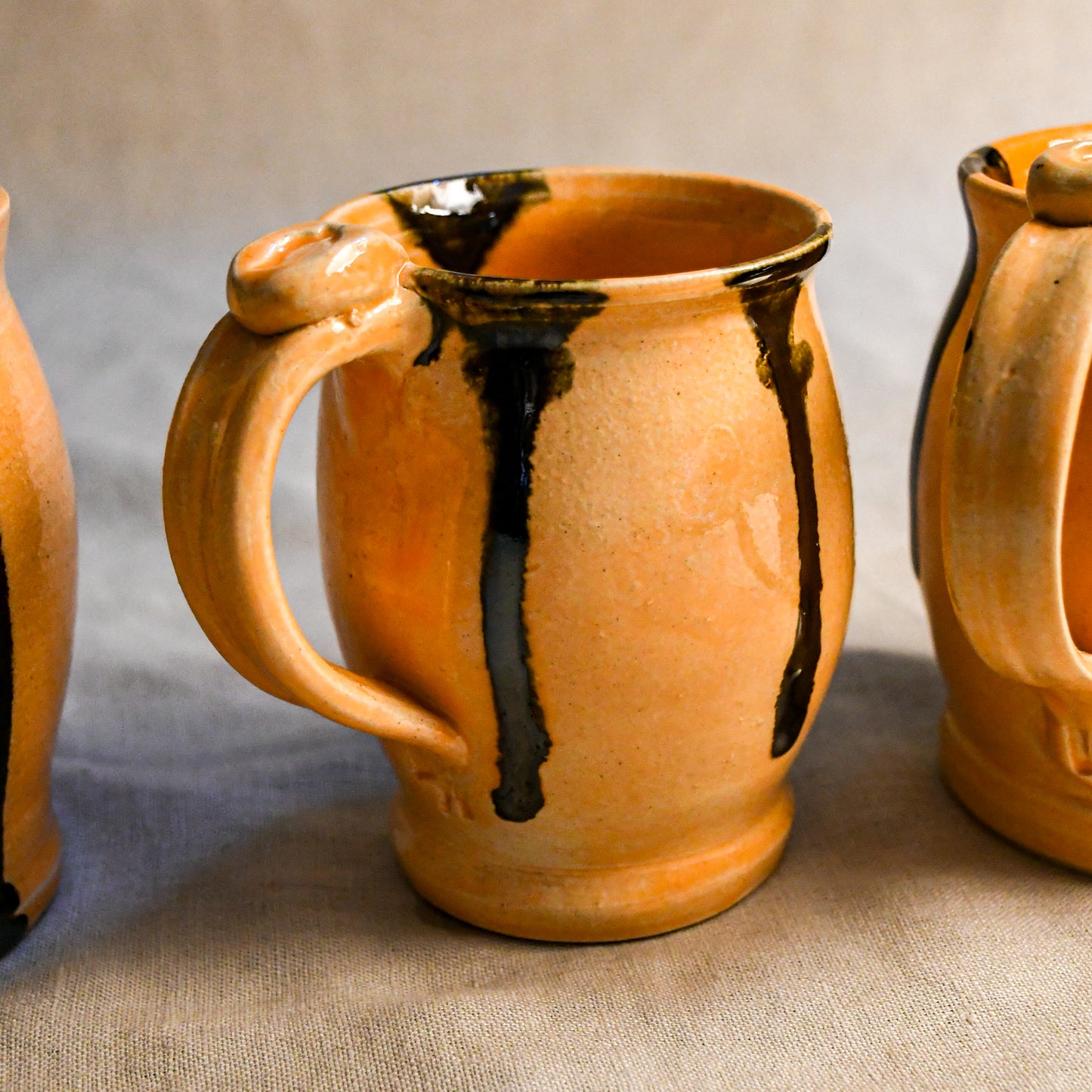 A handmade ceramic mug with a warm orange-brown glaze and dark brown to black dripping accents, shown on a neutral fabric background.