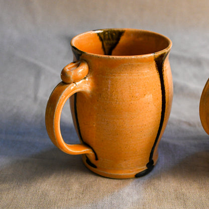 A handmade ceramic mug with a warm orange glaze and dark brown/black drips, shown on a neutral gray fabric background.