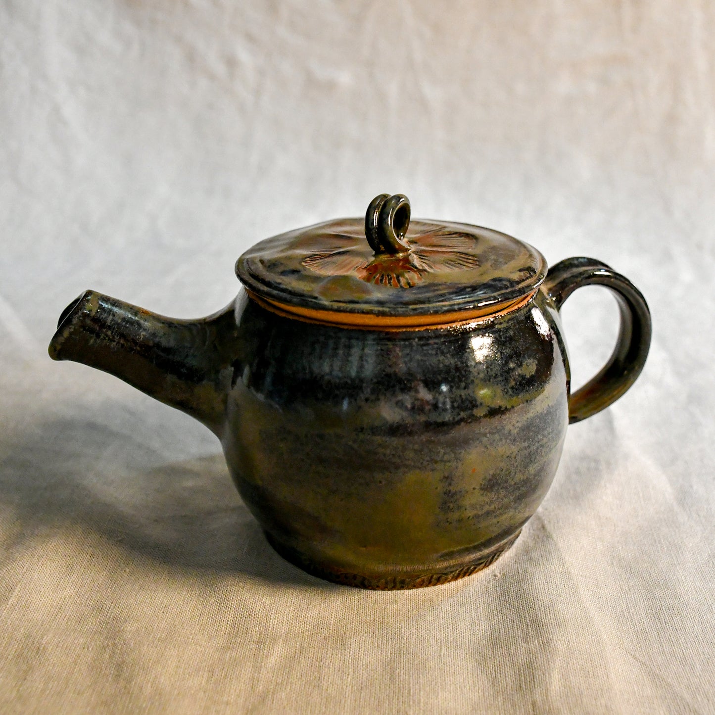 Handmade ceramic teapot in Ancient Jasper glaze featuring a rounded body, curved spout, loop handle, and floral carved lid with rich tones of black, olive, bronze, and rust.
