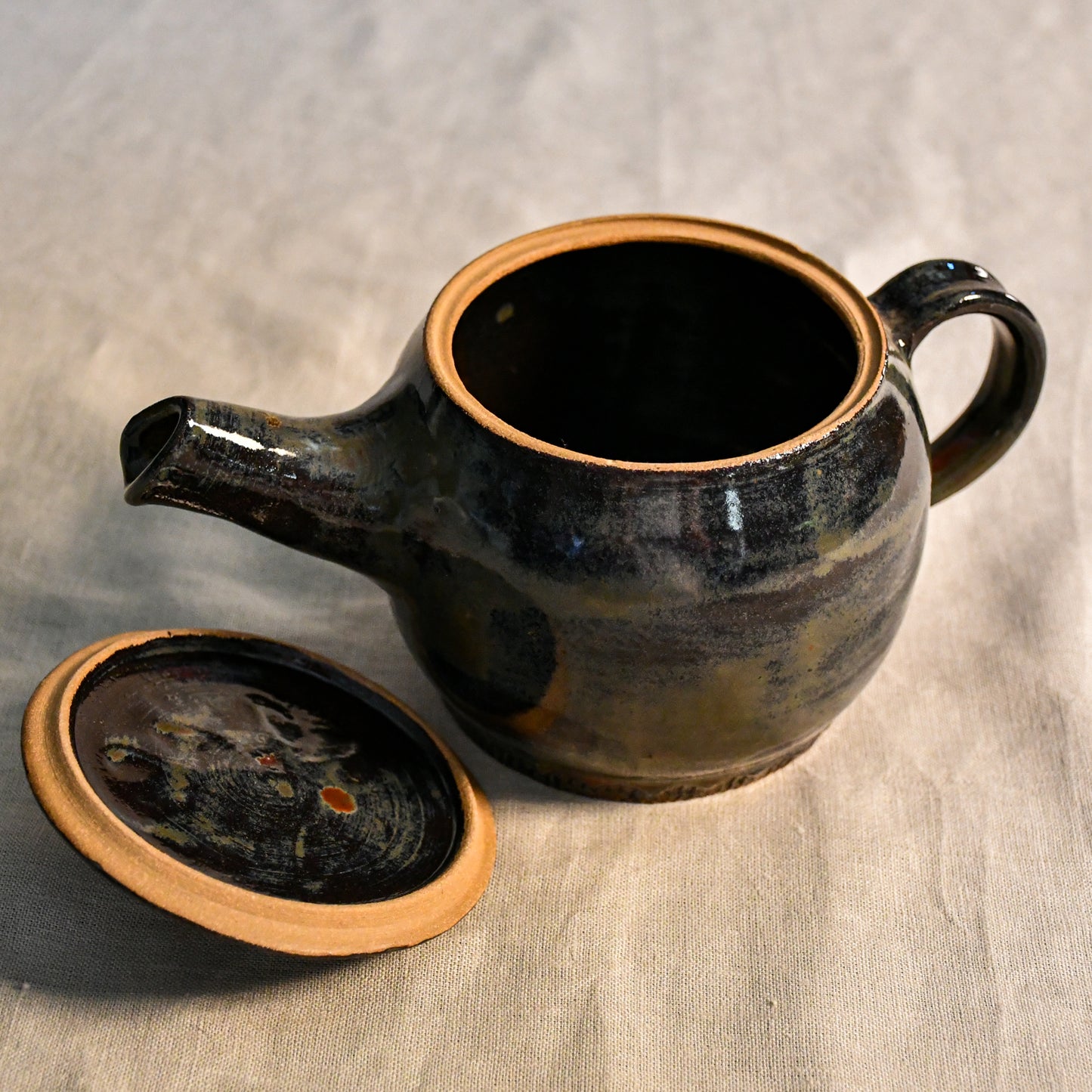 Handmade ceramic teapot in Ancient Jasper glaze featuring a rounded body, curved spout, loop handle, and floral carved lid with rich tones of black, olive, bronze, and rust.