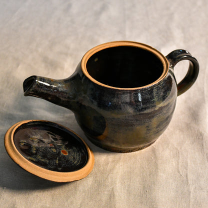 Handmade ceramic teapot in Ancient Jasper glaze featuring a rounded body, curved spout, loop handle, and floral carved lid with rich tones of black, olive, bronze, and rust.
