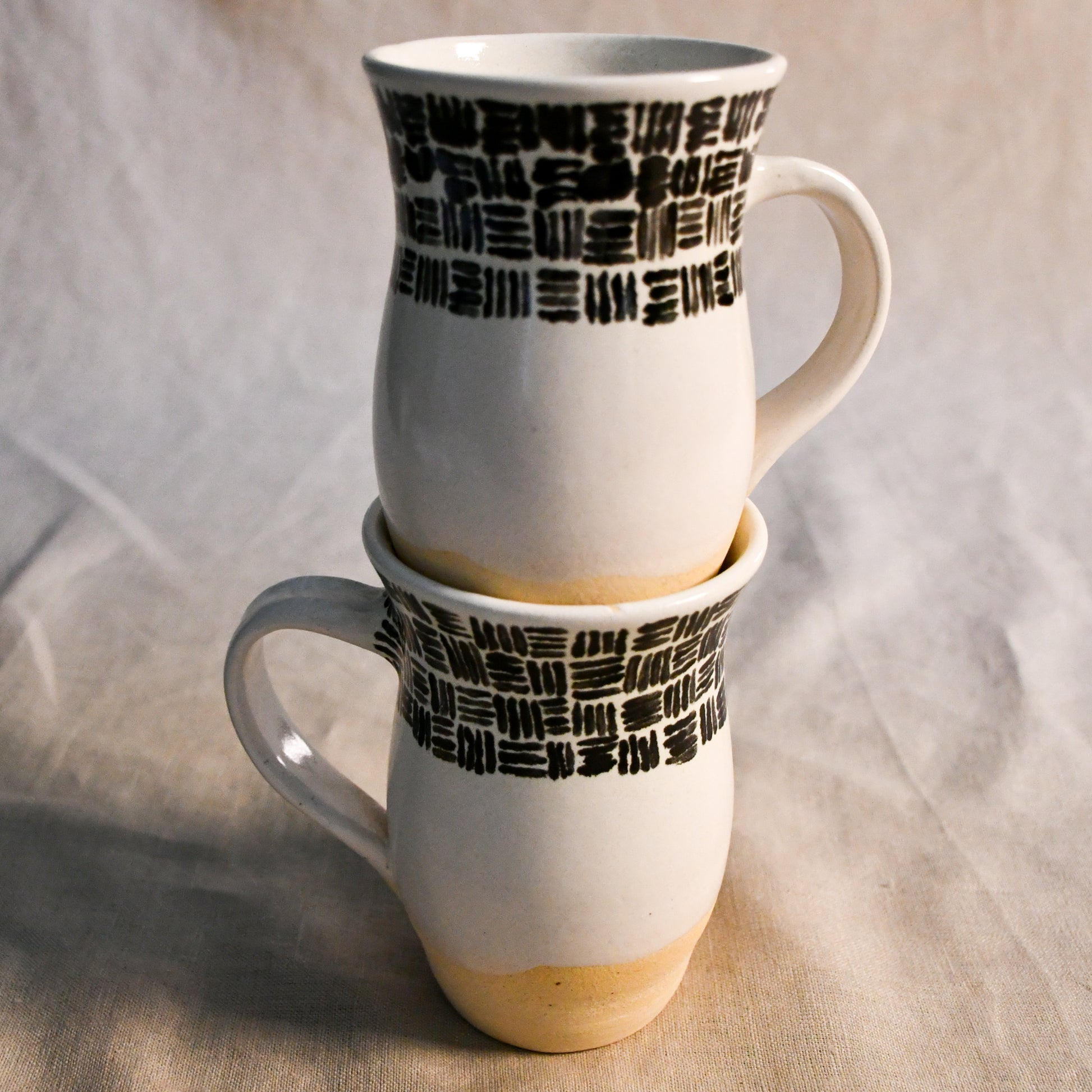 Set of two handmade stoneware mugs by Leigh Art featuring black geometric brushstroke designs, creamy white glaze, and exposed buff clay bases.