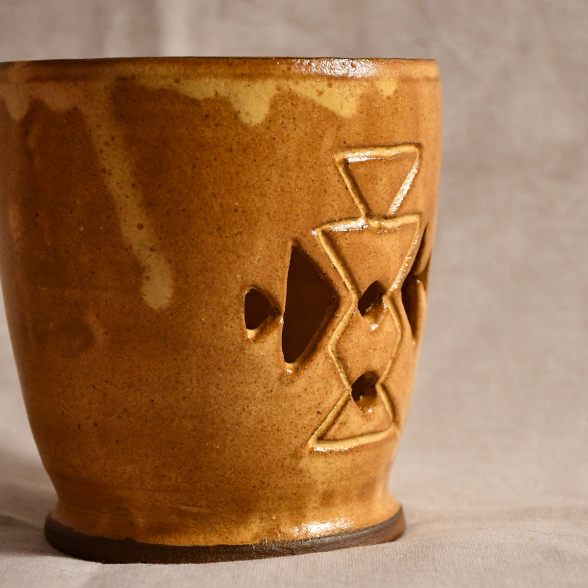 A handmade ceramic candle holder with a warm brown and gold glaze, featuring geometric cutouts on the side, set against a neutral beige fabric background.