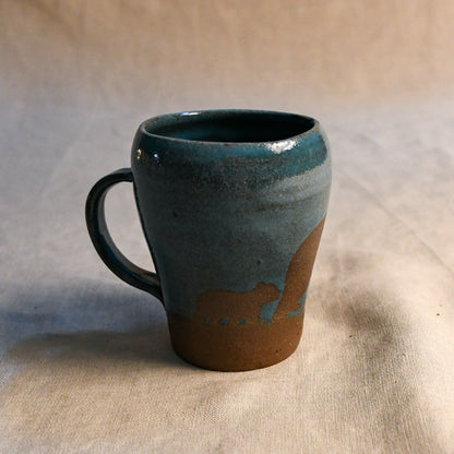 A handmade ceramic mug with a teal blue glaze and a brown unglazed base, shown on a neutral beige fabric background.