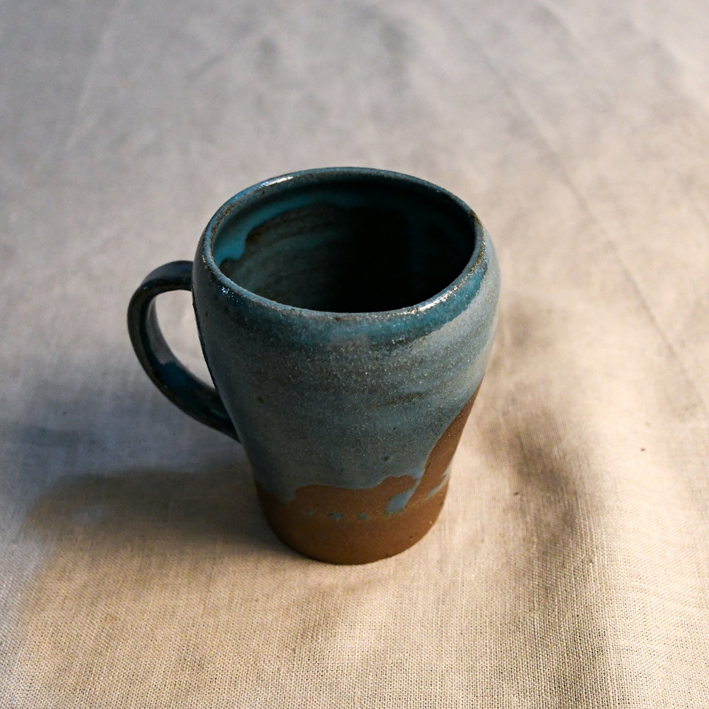 A handmade ceramic mug with a rustic handle, featuring a blue-green glazed finish on the upper body and a natural brown clay base, set on a textured beige linen fabric.