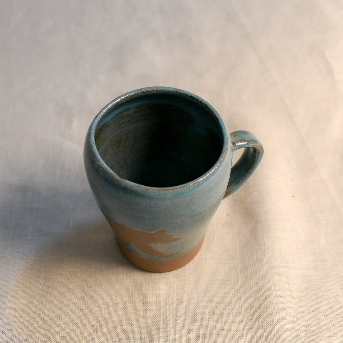 A handmade ceramic mug with a blue-green glazed finish and a natural brown clay base, shown on a light beige textured fabric background.