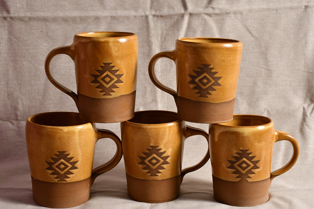 Five ceramic mugs with a rustic two-tone design, featuring a warm ochre upper half and a brown lower half, decorated with a dark brown geometric diamond pattern. They are arranged on a neutral, textured beige fabric backdrop.