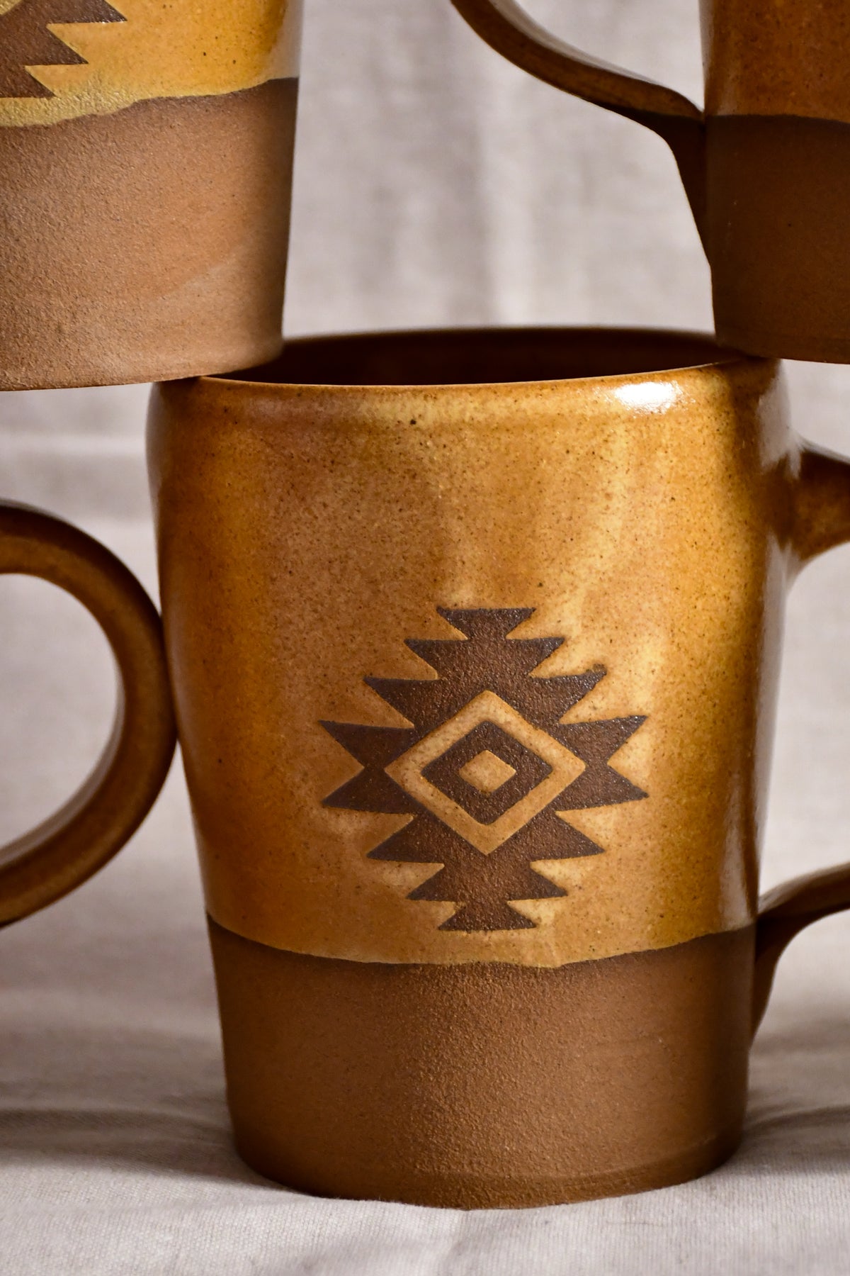 A close-up of a ceramic mug with a Southwestern geometric pattern, featuring a textured metallic gold upper section and a matte brown lower section.