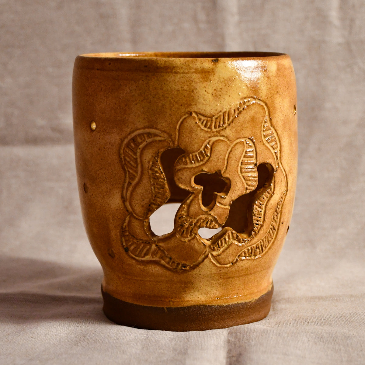 A handmade ceramic candle holder with a warm brown and gold glaze, featuring a floral cutout design on the front and a dark brown base, set against a neutral fabric backdrop.