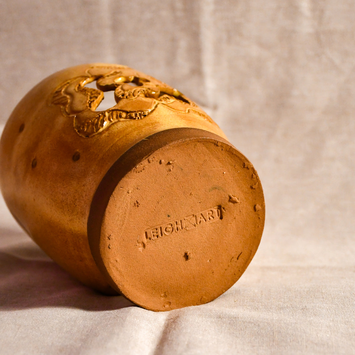 A yellow-colored ceramic jar with a carved floral design on the side, shown lying on its circular base against a neutral fabric backdrop. The brand name 'LEIGH ART' is embossed on the bottom.