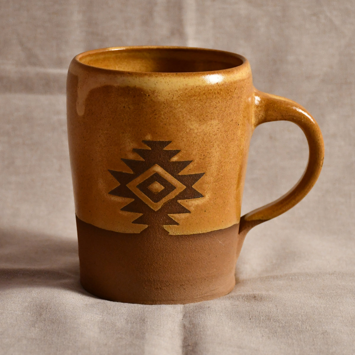 A handmade ceramic mug with a rustic southwestern design, featuring a two-tone glazed finish in warm brown and tan with a dark brown geometric diamond pattern on the front, set against a neutral draped fabric background.