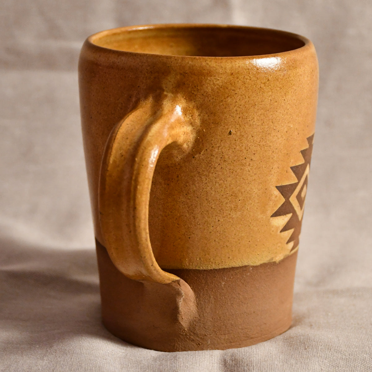 A handmade ceramic mug with a rustic, textured finish, featuring a glossy ochre-brown glaze on the upper body and a matte terracotta-brown base. It has a thick, sculpted handle and a geometric cutout pattern on the side, set against a neutral fabric backdrop.