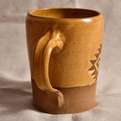A handmade ceramic mug with a rustic, textured finish, featuring a glossy ochre-brown glaze on the upper body and a matte terracotta-brown base. It has a thick, sculpted handle and a geometric cutout pattern on the side, set against a neutral fabric backdrop.