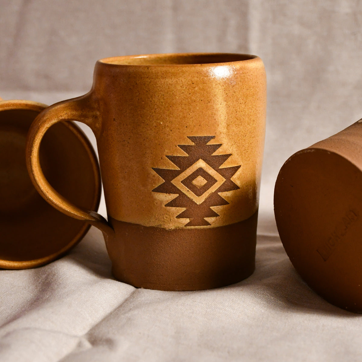 A ceramic mug with a warm brown and tan gradient glaze, featuring a geometric diamond pattern in the center. A matching lid is partially visible on the left and another cropped view of the mug sits on the right, all resting on a neutral fabric surface.