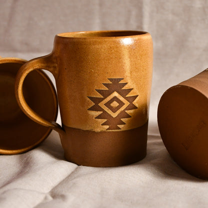 A ceramic mug with a warm brown and tan gradient glaze, featuring a geometric diamond pattern in the center. A matching lid is partially visible on the left and another cropped view of the mug sits on the right, all resting on a neutral fabric surface.