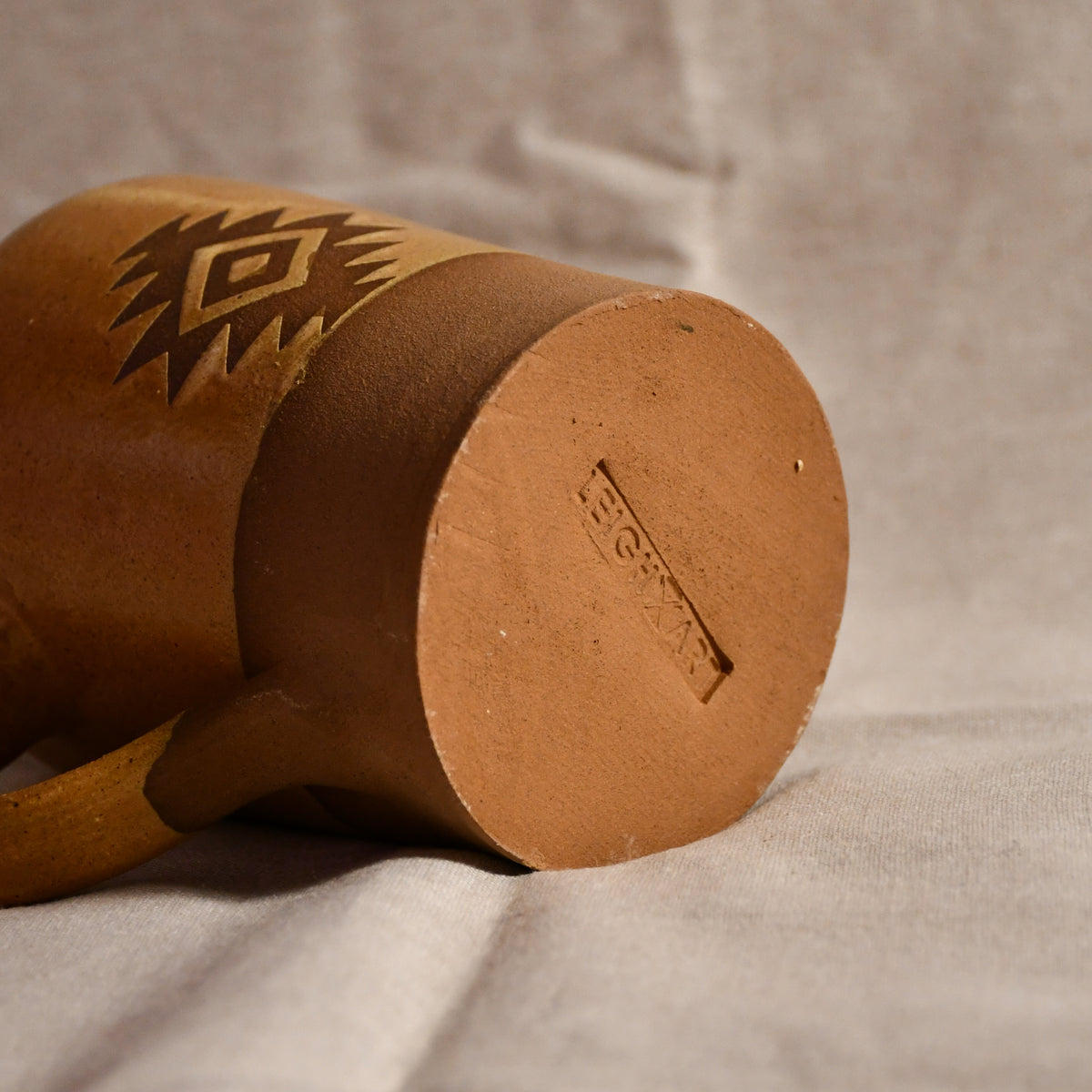 A close-up of a brown ceramic mug showing its bottom edge with a stamped maker's mark and a carved geometric pattern on the side, resting on a textured light beige fabric background.