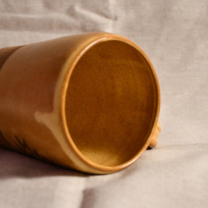 A close-up of a handmade ceramic mug with a warm brown speckled glaze, lying on its side on a textured light gray fabric background.