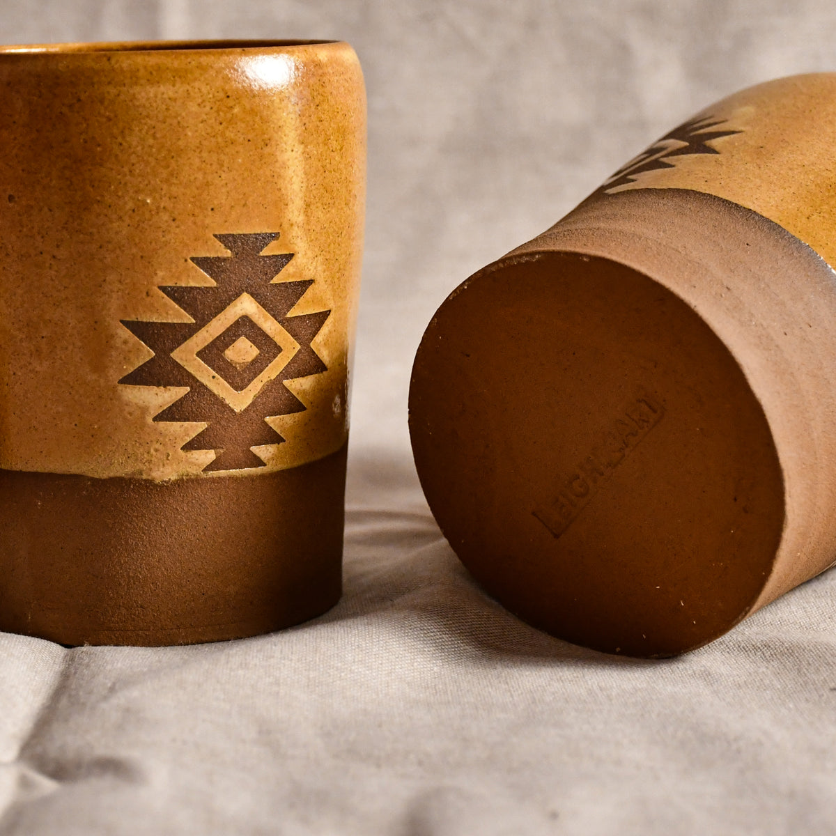 Two rustic ceramic tumblers with a Southwestern geometric pattern, one standing upright and the other lying on its side to show the bottom, on a textured light gray fabric background.