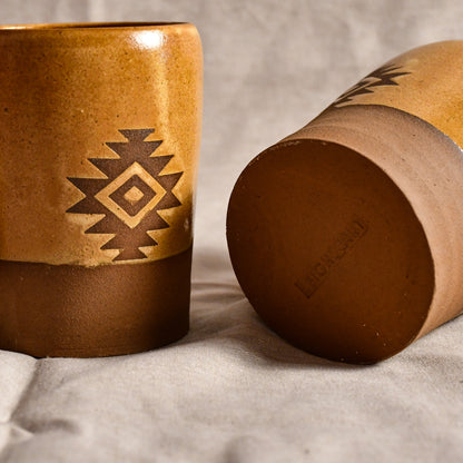 Two rustic ceramic tumblers with a Southwestern geometric pattern, one standing upright and the other lying on its side to show the bottom, on a textured light gray fabric background.