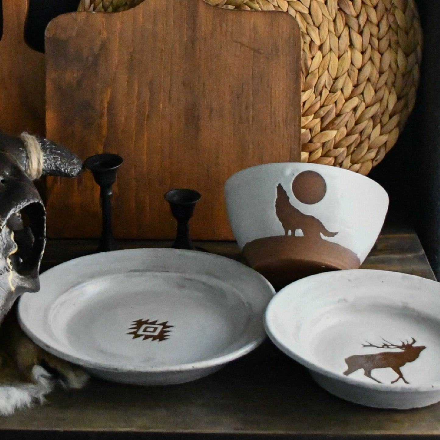 Three handmade ceramic dishes with rustic brown painted designs, including a wolf, a deer, and a geometric sun symbol, displayed on a dark wooden shelf alongside a wooden cutting board, woven basket, candlesticks, and a skull.
