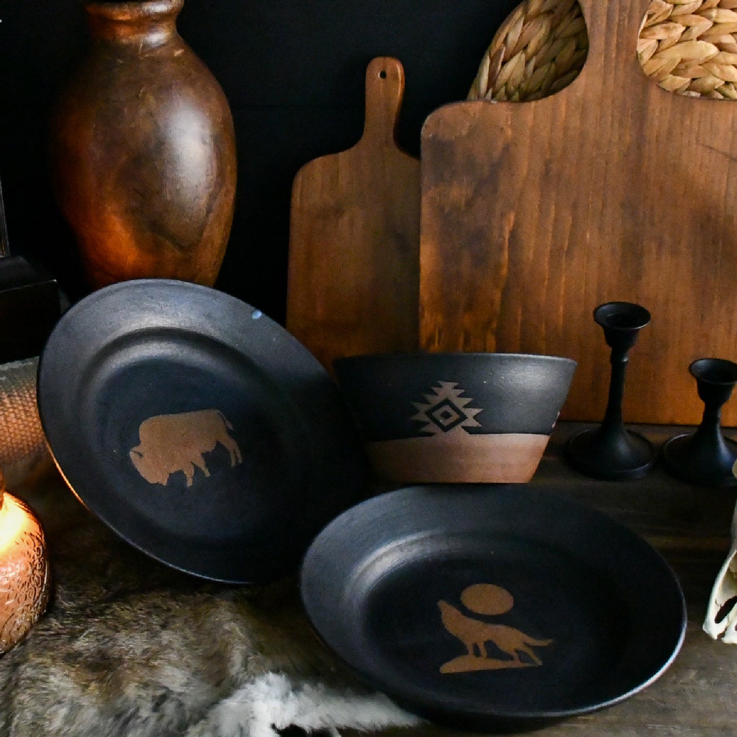A rustic set of dark ceramic tableware, including a plate, bowl, and small dish, decorated with tan Native American-style geometric and animal motifs, displayed on a dark surface with wooden cutting boards and vases in the background.