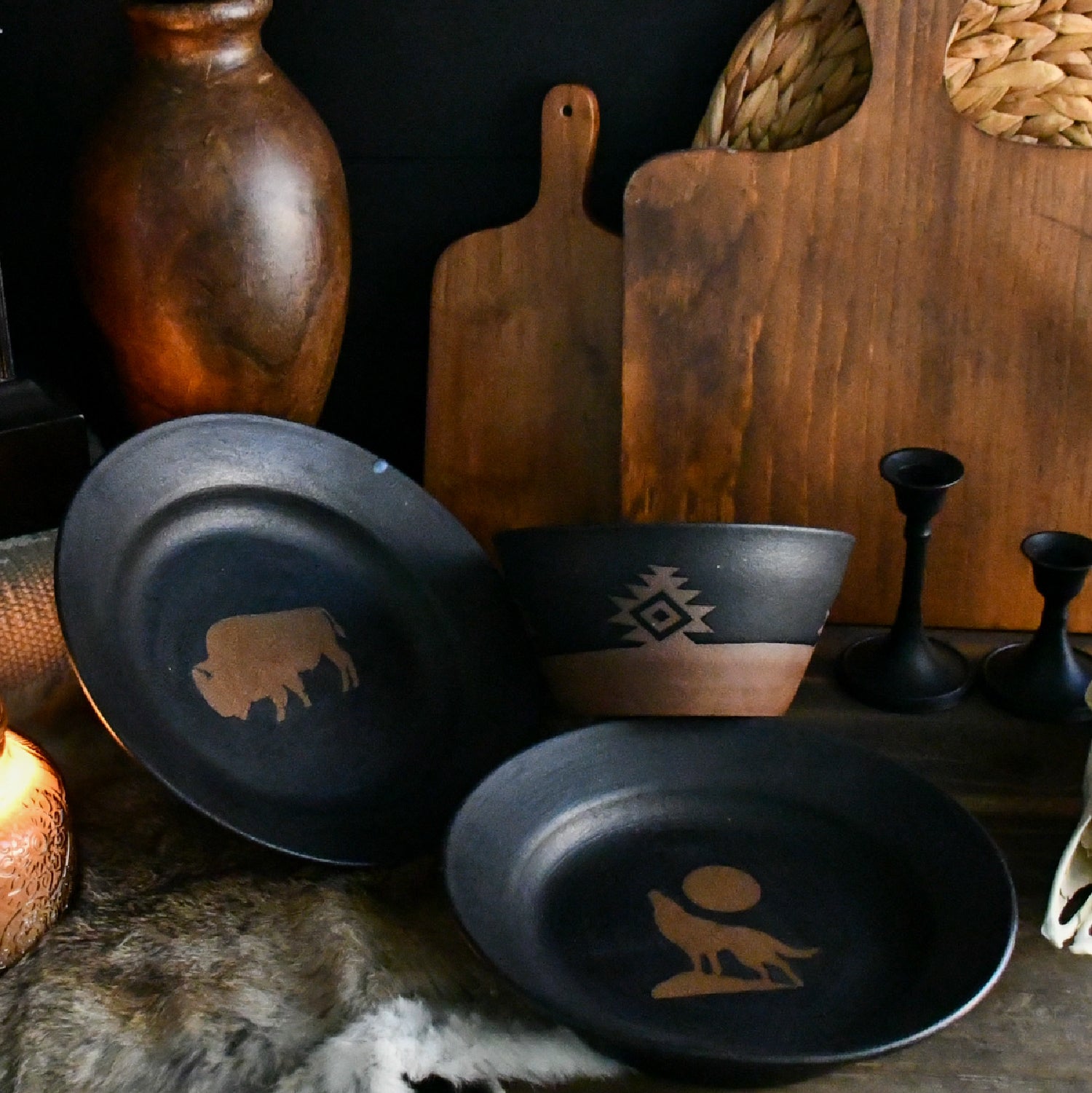A rustic set of dark ceramic tableware, including a plate, bowl, and small dish, decorated with tan Native American-style geometric and animal motifs, displayed on a dark surface with wooden cutting boards and vases in the background.