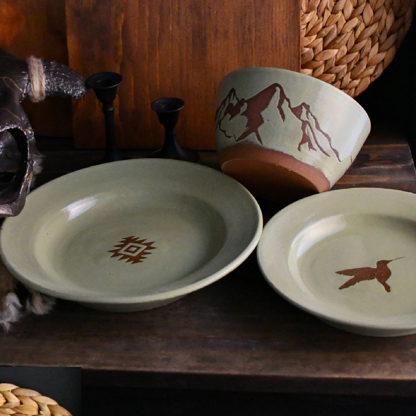 Three handmade ceramic tableware pieces displayed on a dark wooden surface: a shallow round plate with a brown geometric symbol, a bowl with a brown mountain design, and a shallow plate with a brown hummingbird. Decorative rustic props including a woven basket, candlesticks, and a dark metal object frame the scene.