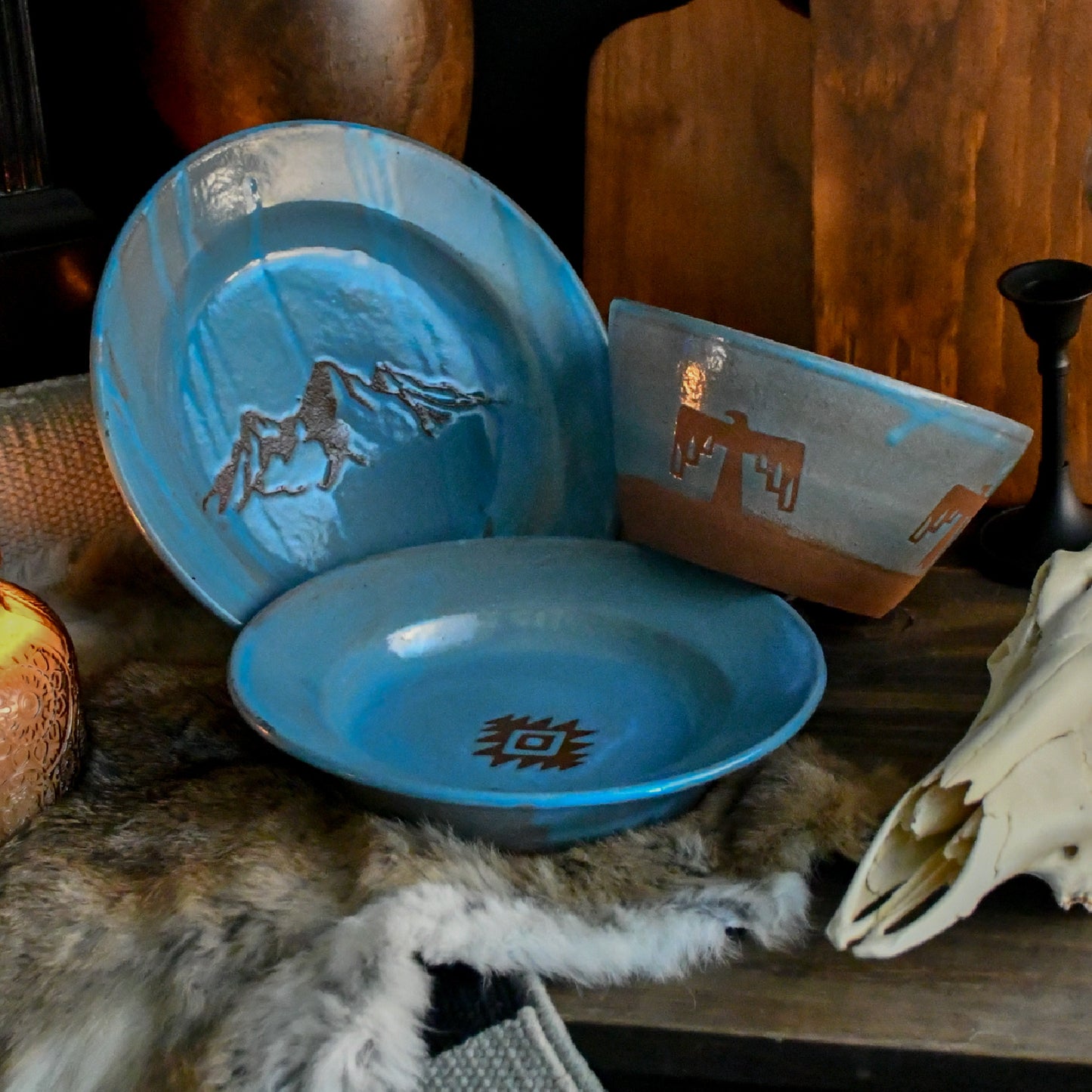 A set of rustic handmade ceramic tableware, including a blue plate with a mountain design, a matching bowl, and a rectangular dish, styled on a dark wooden table with fur, a skull, and candlesticks.
