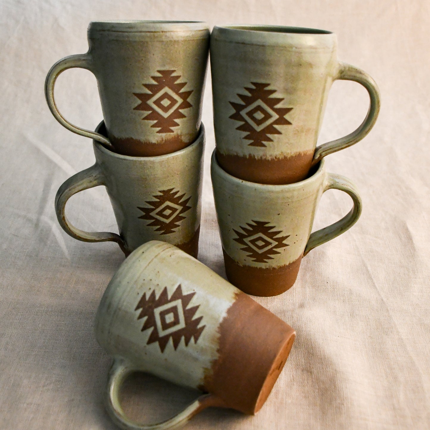 Five handmade Wild Rojo pottery mugs featuring an Aztec-inspired design in earthy green glaze, stacked together to showcase the coordinating mug pack.